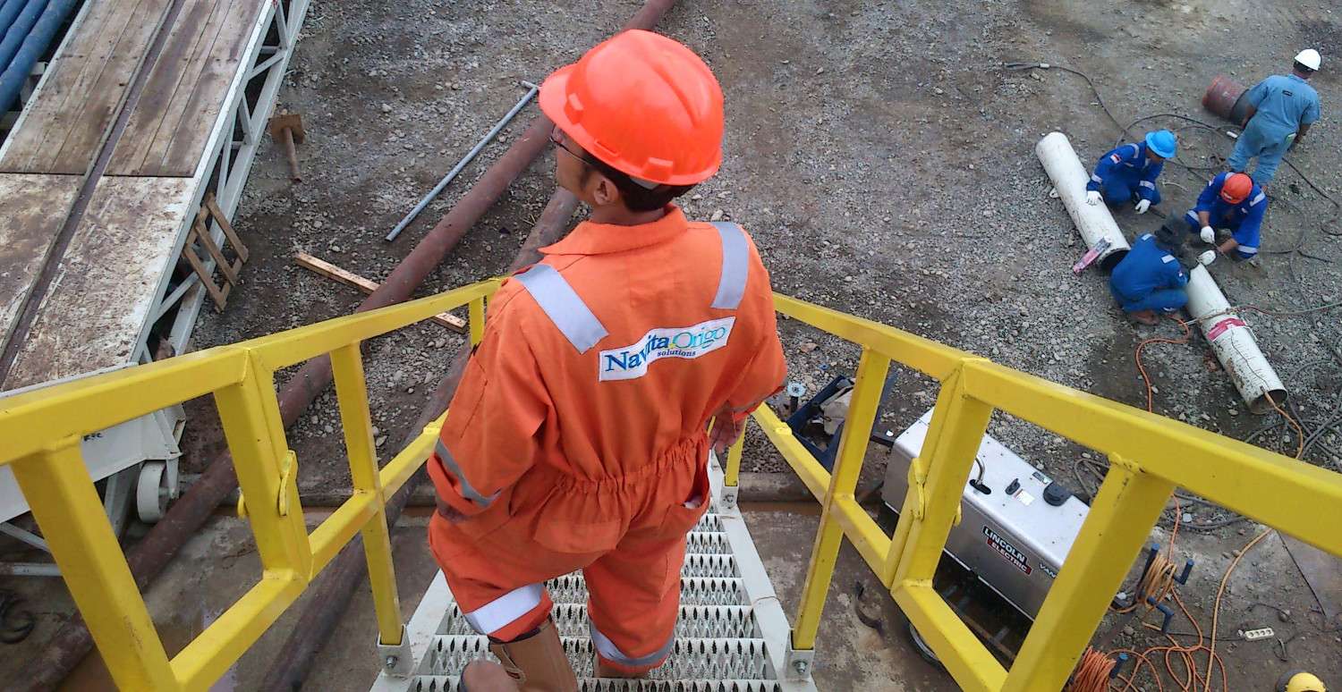 A Navita Origo field engineer in branded coverall and hard hat on the wellsite stairway, with the crew working on casing below